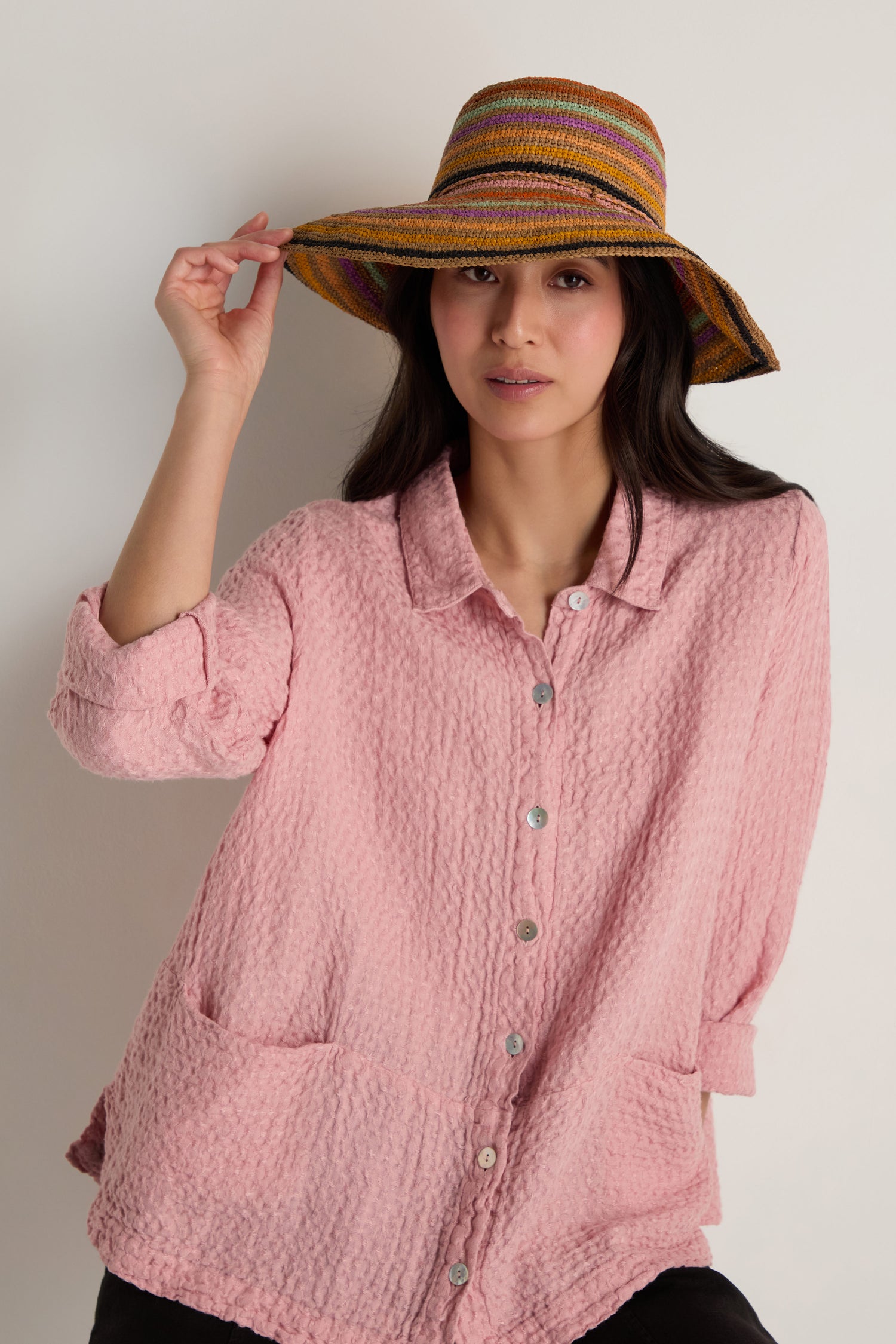 A woman models the Handwoven Raffia Rainbow Stripe Hat and a textured pink button-up shirt against a plain light background.
