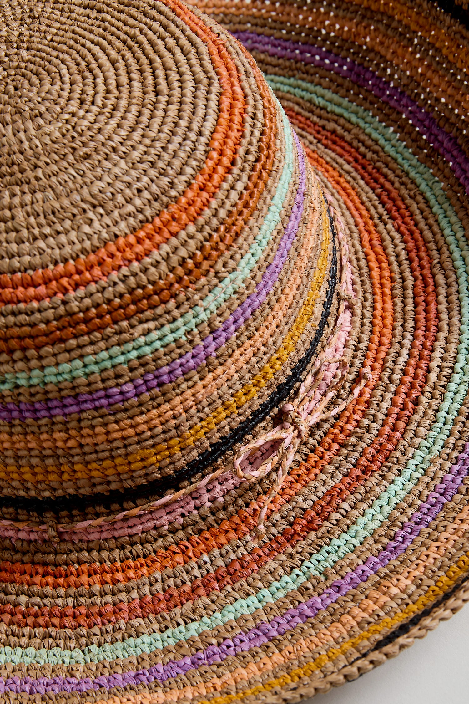 Close-up of the Handwoven Raffia Rainbow Stripe Hat, crafted in Madagascar with vibrant orange, green, purple, pink, and black stripes, a wide brim for style, and a side bow detail.