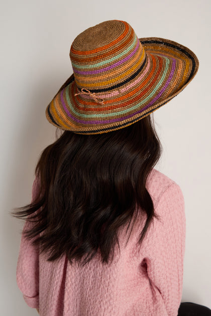 A person with long dark hair faces away from the camera, wearing the Handwoven Raffia Rainbow Stripe Hat featuring a wide brim and a textured pink top, set against a plain background.