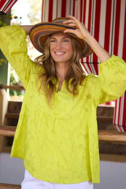 A woman in a yellow textured blouse and white pants smiles while holding the Handwoven Raffia Rainbow Stripe Hat, featuring a colorful wide brim, standing before red-and-white striped outdoor curtains.