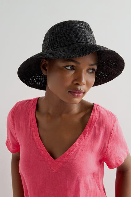 A person in a pink V-neck shirt and a Handwoven Raffia Hat looks to the side against a light background. The hat, made with Madagascar weaving techniques, offers an elegant and breathable touch to the ensemble.