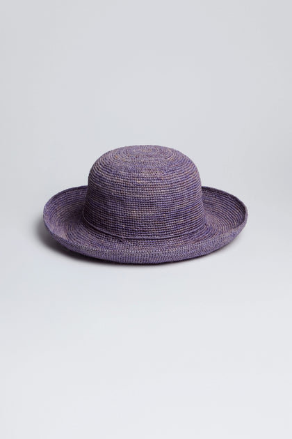 The Handwoven Raffia Hat, crafted in Madagascar, boasts a wide brim and a lovely purple hue, sitting elegantly on any surface.