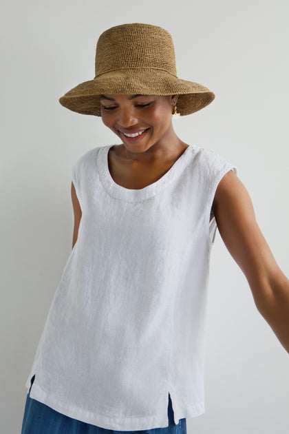 Wearing a Handwoven Raffia Hat, a sleeveless white top, and blue pants, a person smiles while glancing down against a plain background.