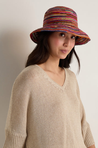 A woman in a beige knit sweater wears the Handwoven Rainbow Raffia Hat, standing against a plain light-colored wall.