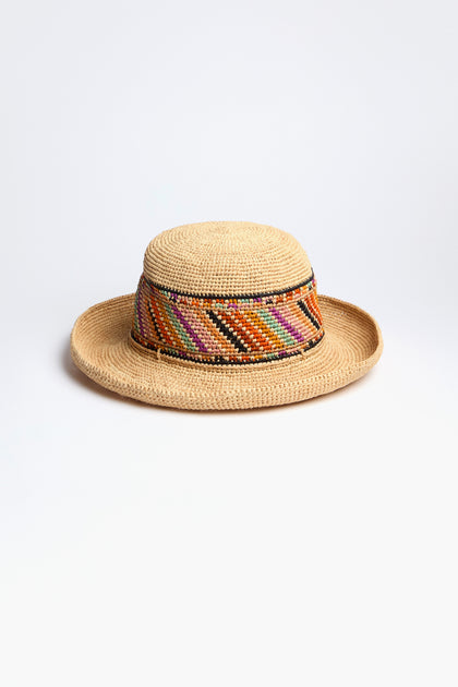 The Handwoven Aztec Raffia Hat is a straw sun hat with a wide brim and features a colorful diagonal striped band in red, orange, and green.