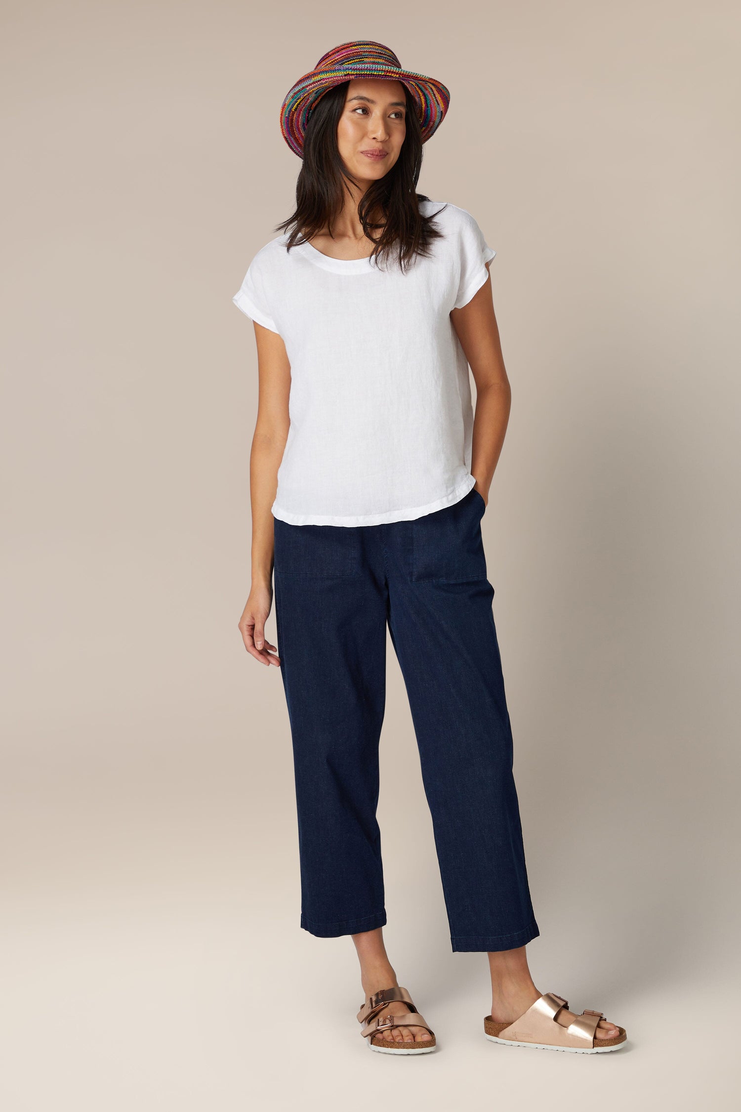 A woman standing in a neutral pose wearing a white t-shirt, blue cropped pants, sandals, and a Woven Rainbow Raffia Hat.