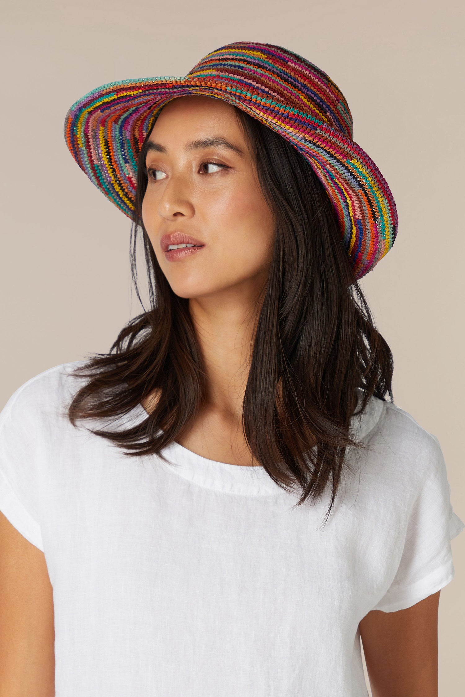 A woman wearing a white shirt and a Woven Rainbow Raffia Hat looks to the side.