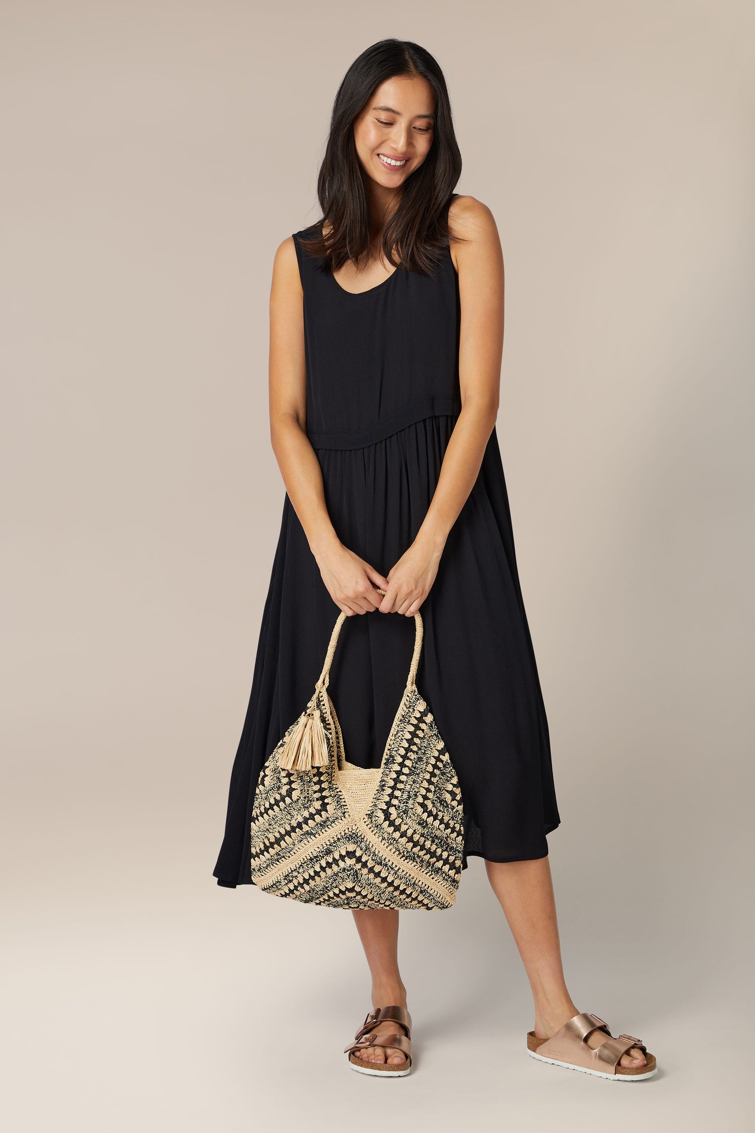 A woman in a black sleeveless dress smiling and holding a Woven Bohemian Raffia Bag.