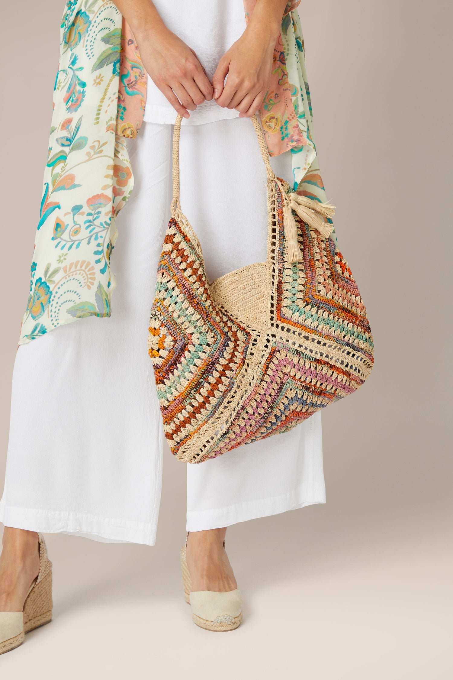 Woman holding a colorful Rabarany Woven Bohemian Raffia Bag while wearing white pants and beige shoes.