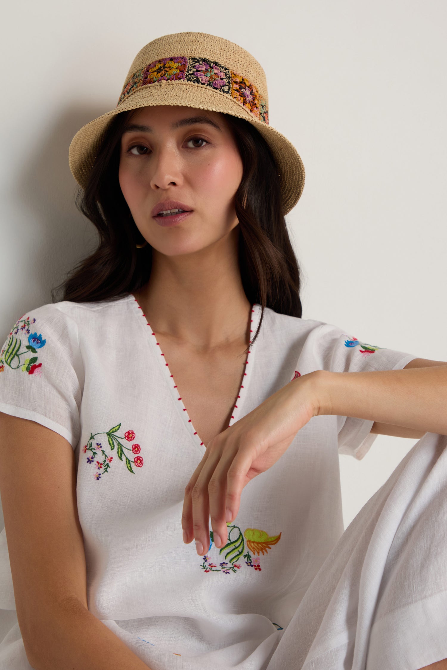 A woman wearing a white embroidered outfit with floral crochet details models the Handwoven Raffia Flower Bucket Hat, seated against a plain white background.