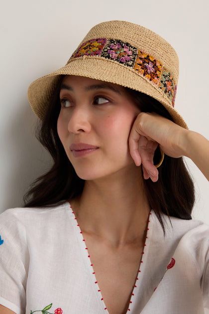 A woman wearing the Handwoven Raffia Flower Bucket Hat, featuring colorful floral crochet from Madagascar, and a white embroidered top rests her chin on her hand, gazing to the side.