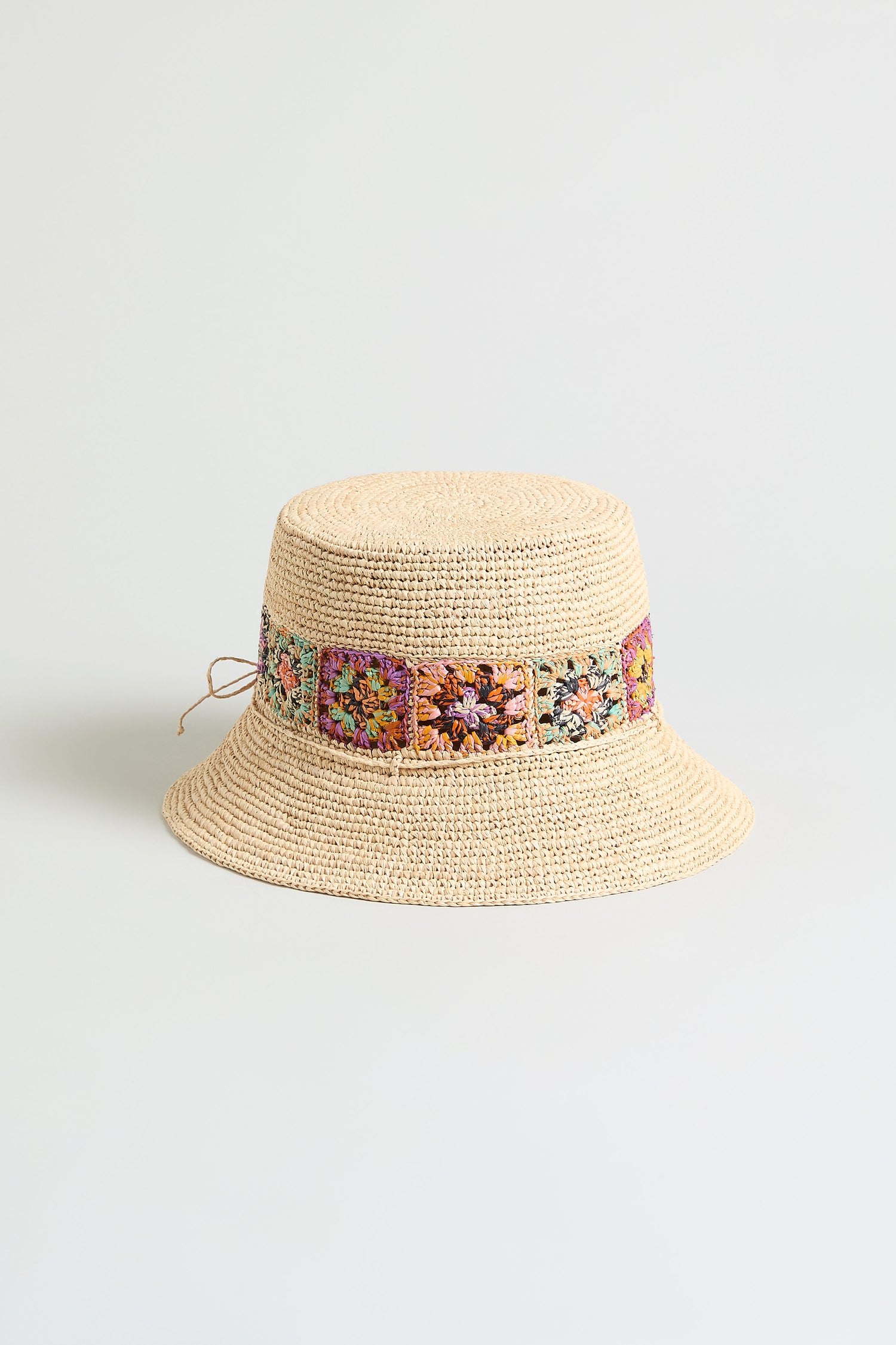 The Handwoven Raffia Flower Bucket Hat, crafted in Madagascar, features beige raffia and colorful floral crochet accents around the crown, shown against a plain light background.