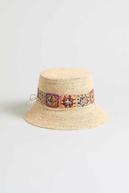 The Handwoven Raffia Flower Bucket Hat, crafted in Madagascar, features beige raffia and colorful floral crochet accents around the crown, shown against a plain light background.