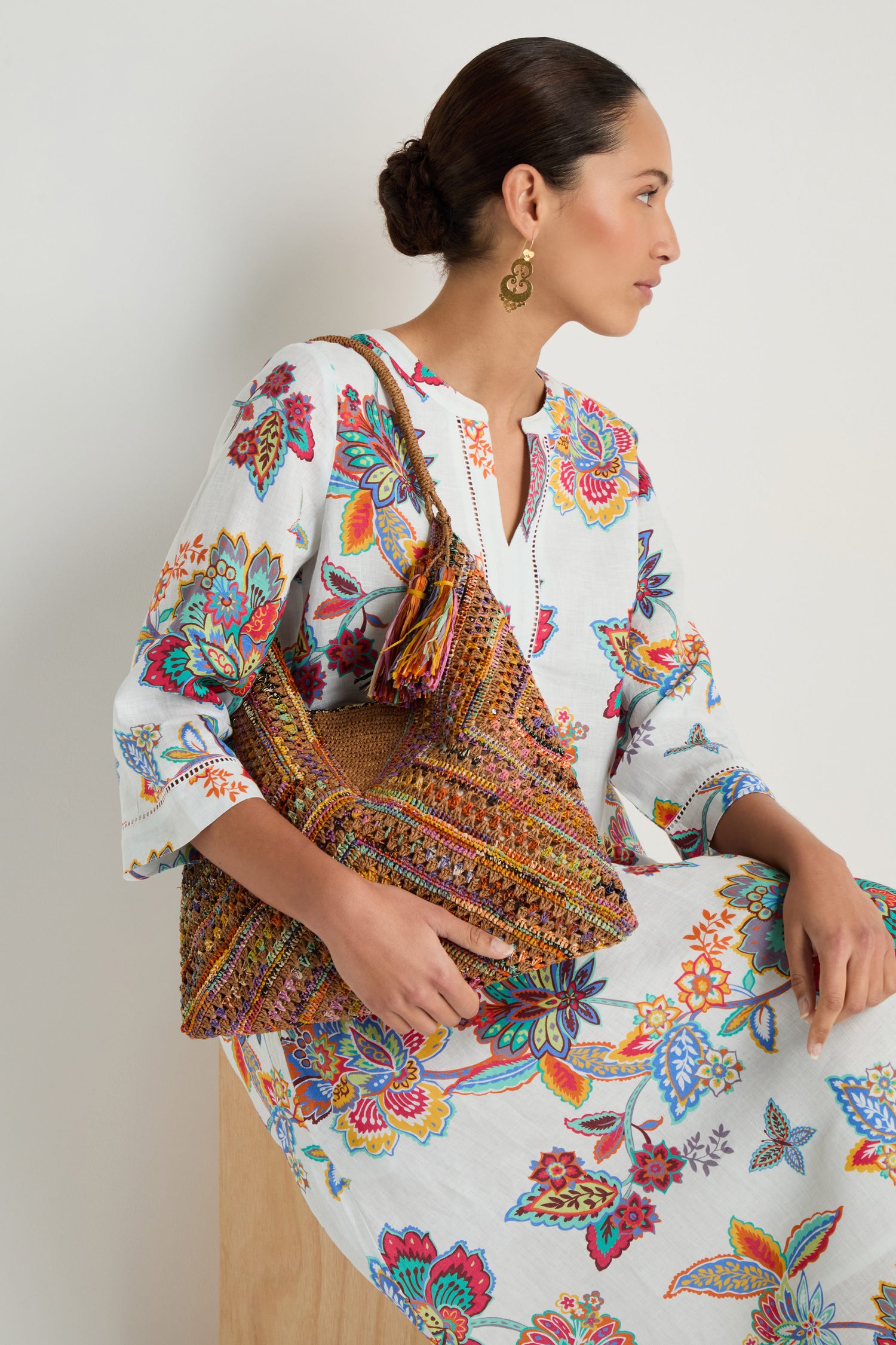 A woman in a white floral dress sits on a wooden block, holding the Handwoven Raffia Harlequin Bag and wearing gold earrings.