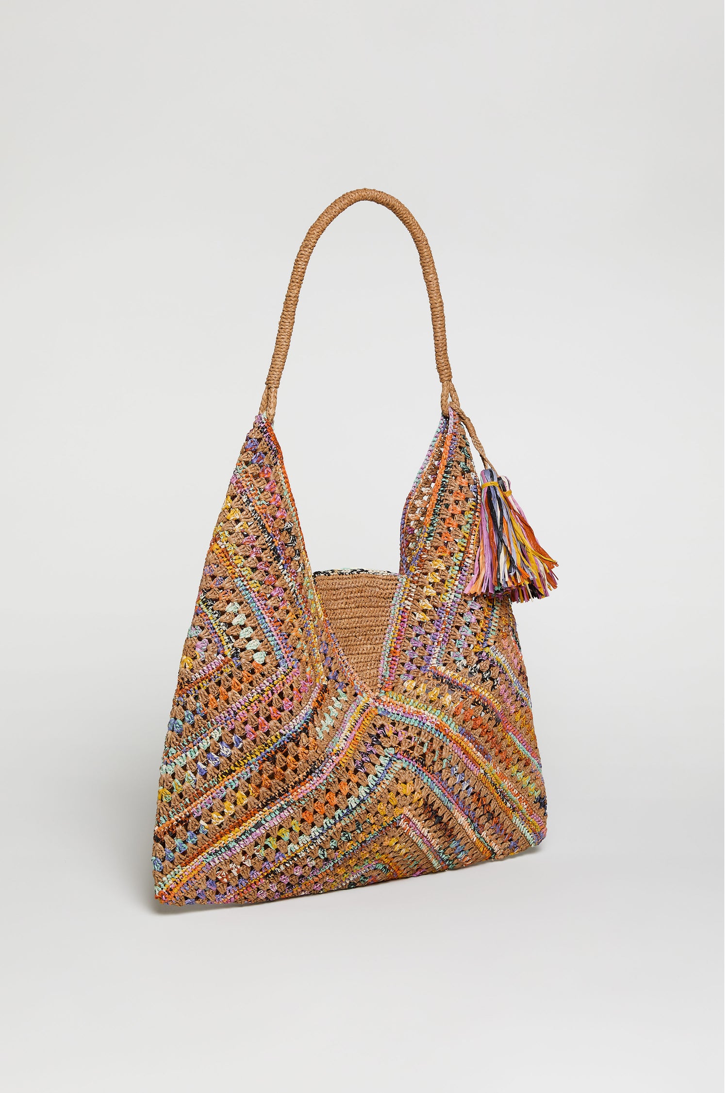 The Handwoven Raffia Harlequin Bag from Madagascar showcases a colorful harlequin pattern, braided handle, and tassel accents, all photographed on a plain white background.