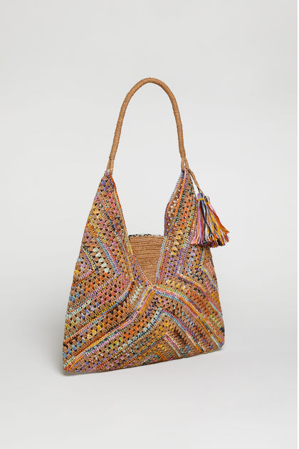 The Handwoven Raffia Harlequin Bag from Madagascar showcases a colorful harlequin pattern, braided handle, and tassel accents, all photographed on a plain white background.