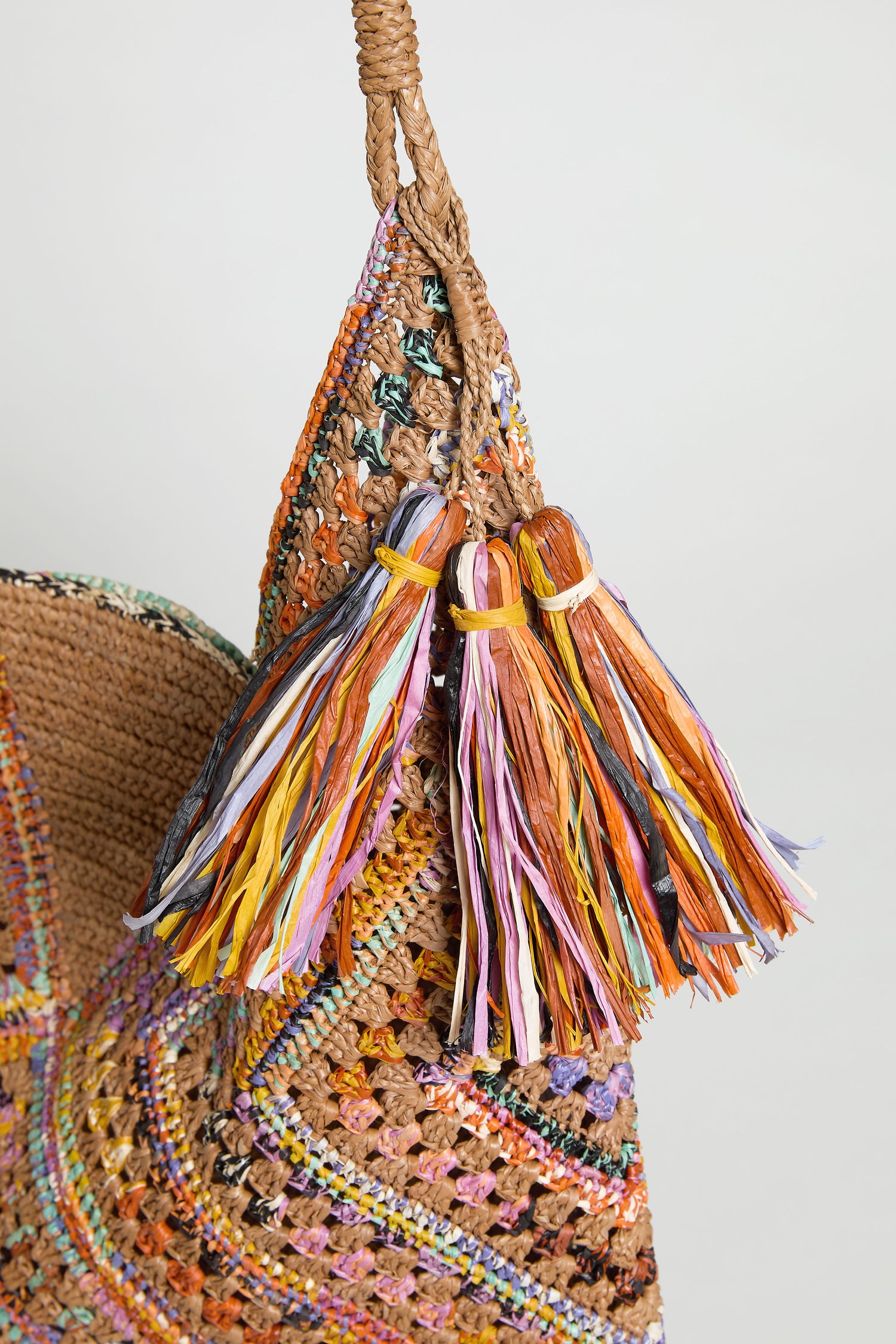 A close-up of the Handwoven Raffia Harlequin Bag from Madagascar, showcasing its multicolored yarn tassels and vibrant decorative stitching.