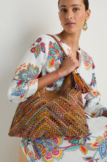 A woman in a white floral dress sits holding the Handwoven Raffia Harlequin Bag from Madagascar, featuring a vibrant harlequin pattern and playful tassel detail.