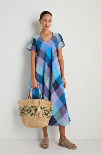 A woman in a blue and purple plaid dress stands against a white wall, holding the Handwoven Raffia Mosaic Bag. She completes her look with sandals and hoop earrings.