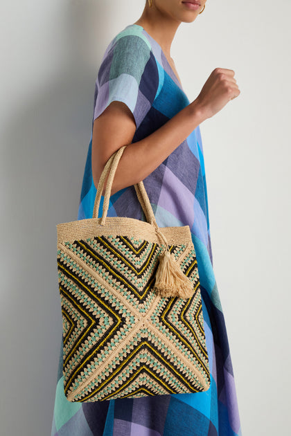 A woman in a blue and purple checkered dress holds the Handwoven Raffia Mosaic Bag, featuring geometric patterns and a tassel, against a plain white wall.