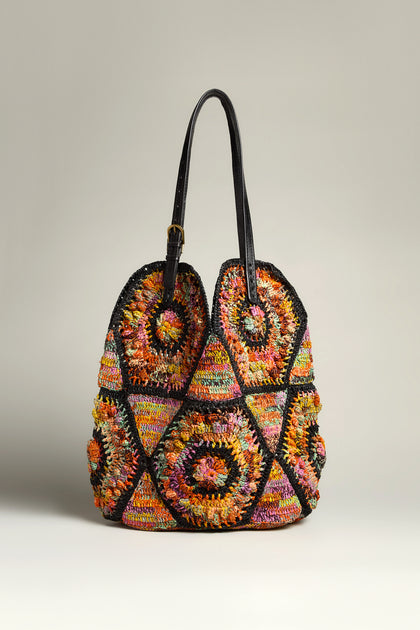 The Handwoven Aztec Raffia Bag features multicolored crochet, circular patterns, black trim, and dual handles. This artisanal piece highlights Madagascar raffia craftsmanship against a neutral background.