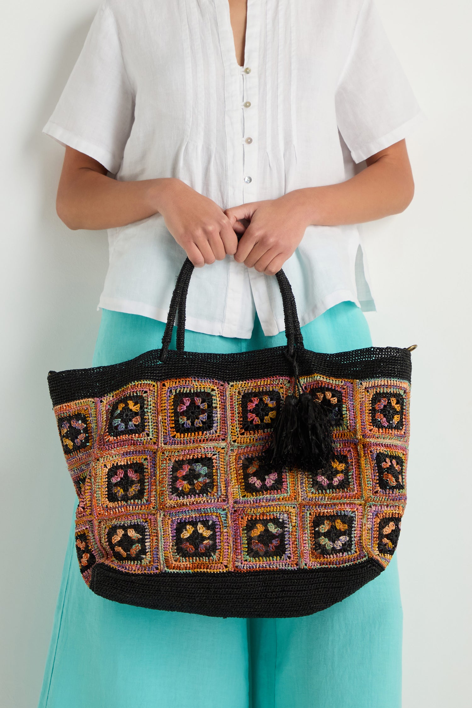 A person wearing a white blouse and turquoise pants holds a large black Handwoven Raffia Crochet Bag from Madagascar, featuring colorful square patterns and finished with a black tassel.