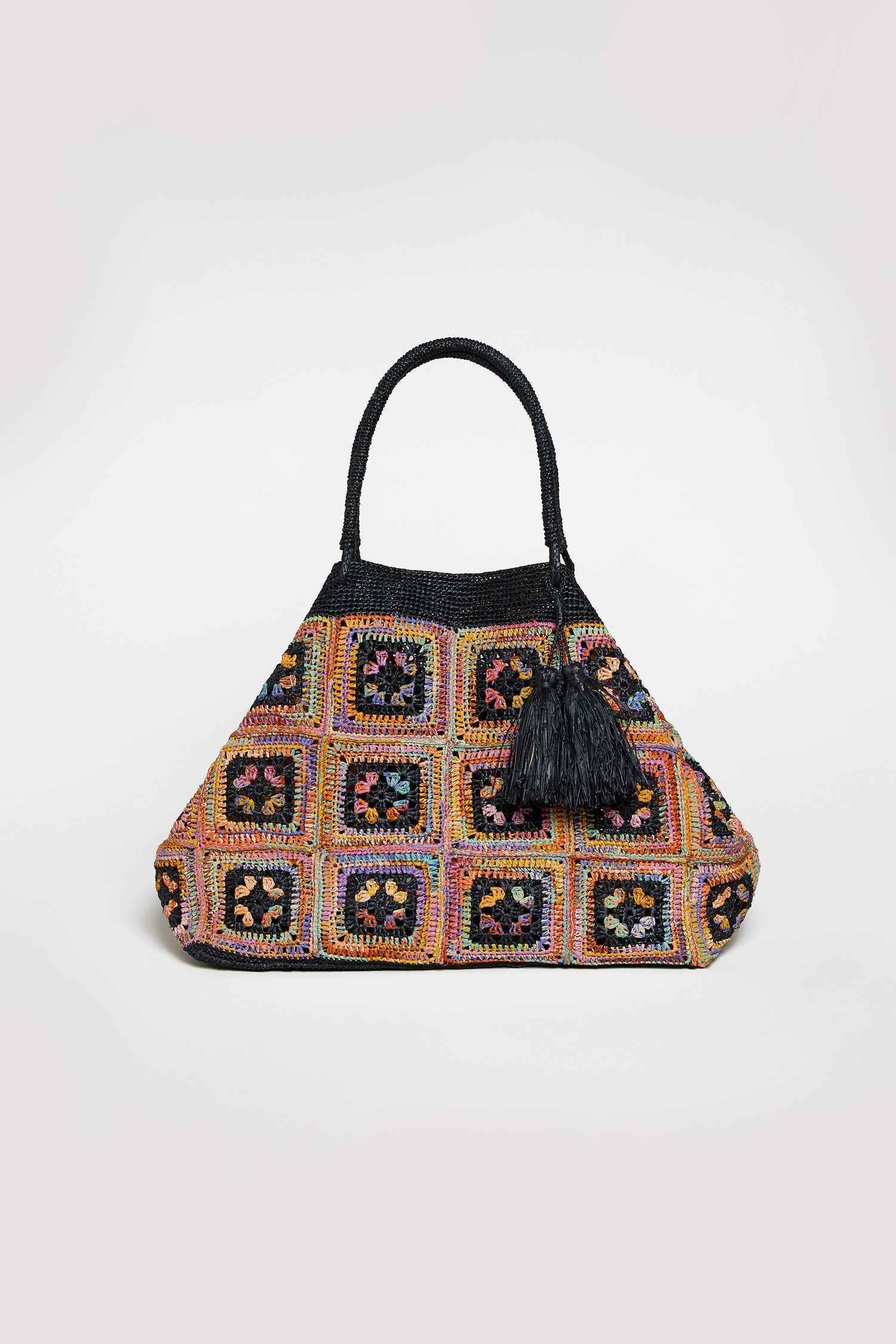 The Handwoven Raffia Crochet Bag features a triangular shape, black handles, a colorful crochet square pattern, and a black tassel by the handles, inspired by Madagascar design, displayed against a simple background.