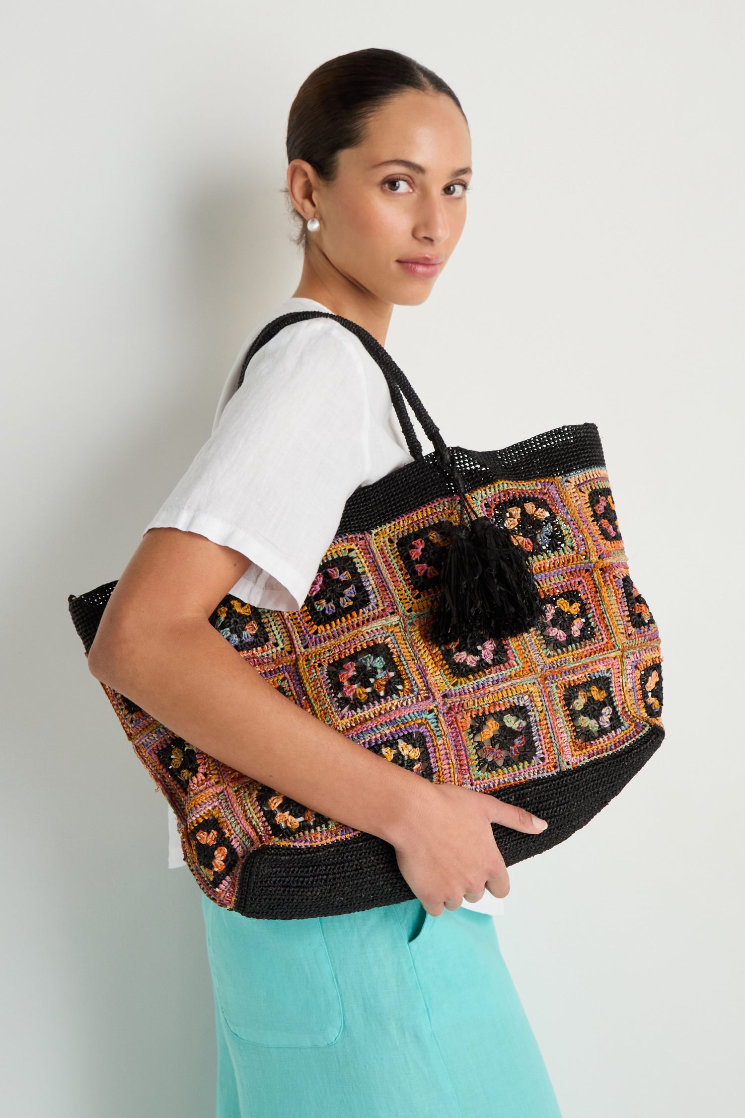 A woman in a white blouse and turquoise skirt carries the Handwoven Raffia Crochet Bag, featuring a black handle and tassel, against a plain background.