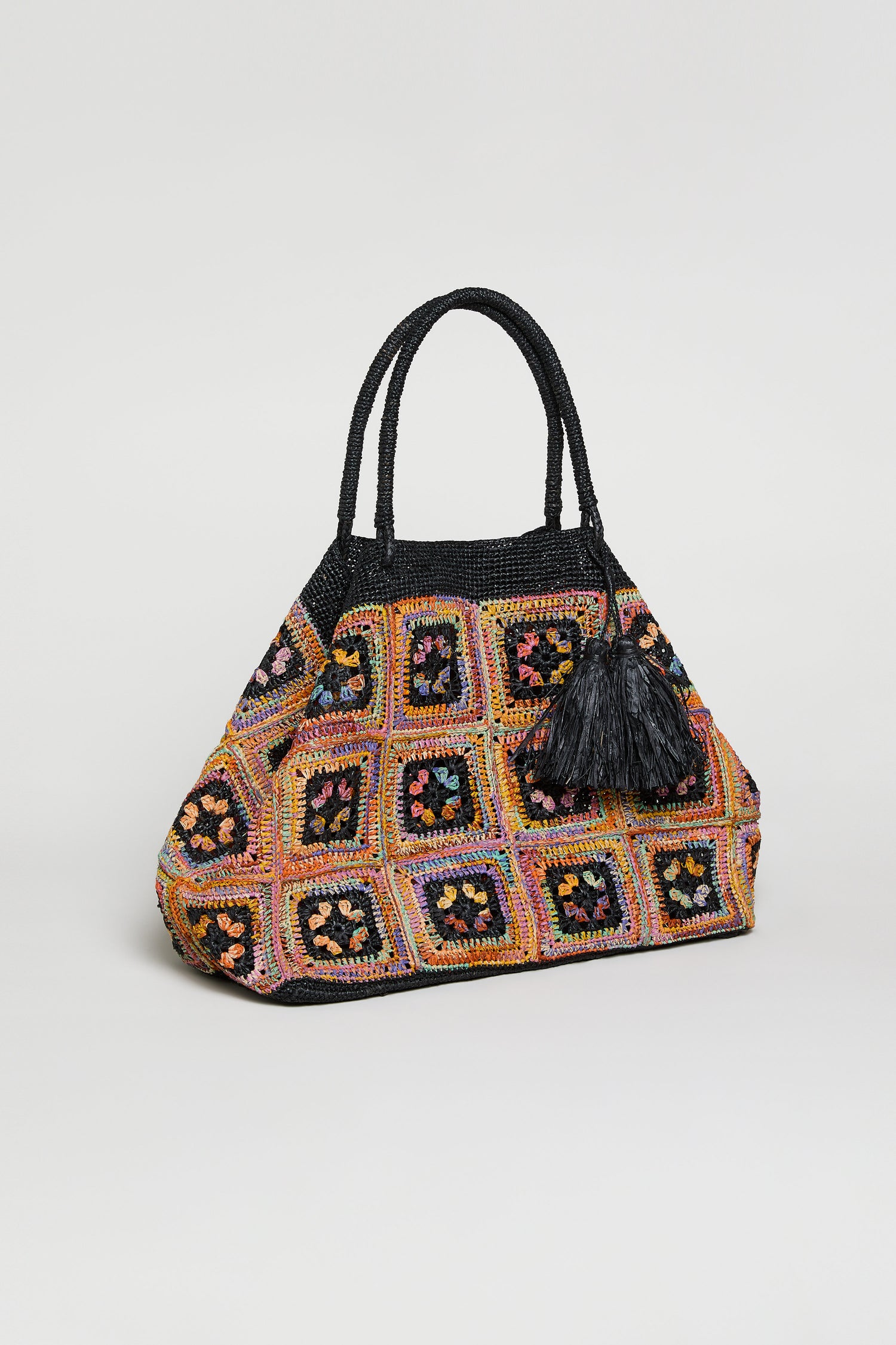 The Handwoven Raffia Crochet Bag from Madagascar features a vibrant multicolored design with black handles, a black tassel, and striking square motifs.