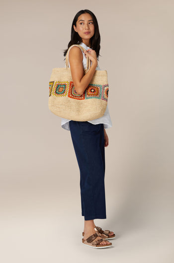 A woman standing to the side and looking at the camera, holding a large patterned Aztec Woven Raffia Bag from Madagascar on her shoulder.