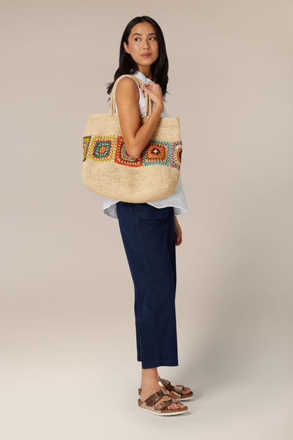 A woman standing to the side and looking at the camera, holding a large patterned Aztec Woven Raffia Bag from Madagascar on her shoulder.