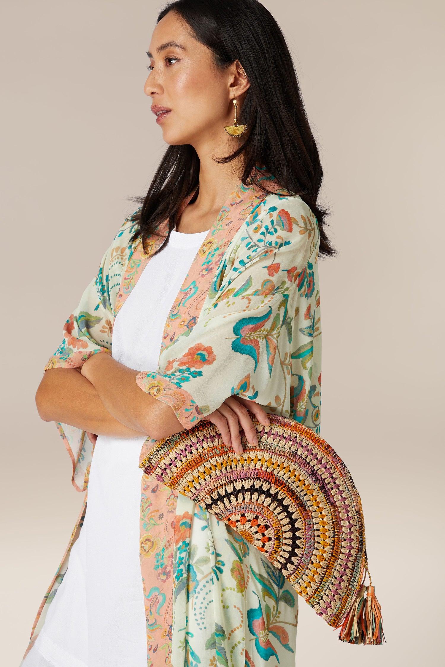 Sentence with Product Name: Woman holding a Woven Half Moon Raffia Clutch and wearing a floral kimono-style cardigan.