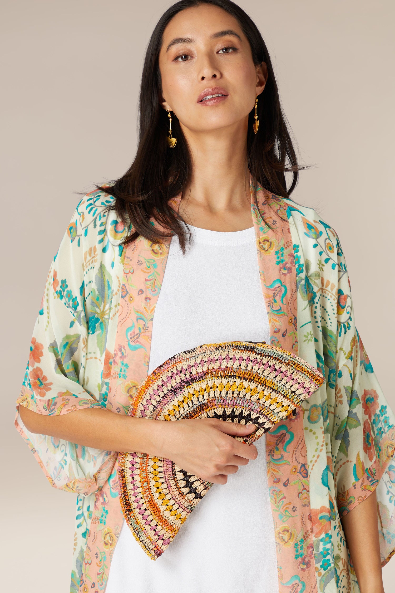 A woman holding a colorful hand fan and wearing a floral kimono over a white top with dangle earrings, accompanied by the Woven Half Moon Raffia Clutch that is Madagascar hand-crafted.