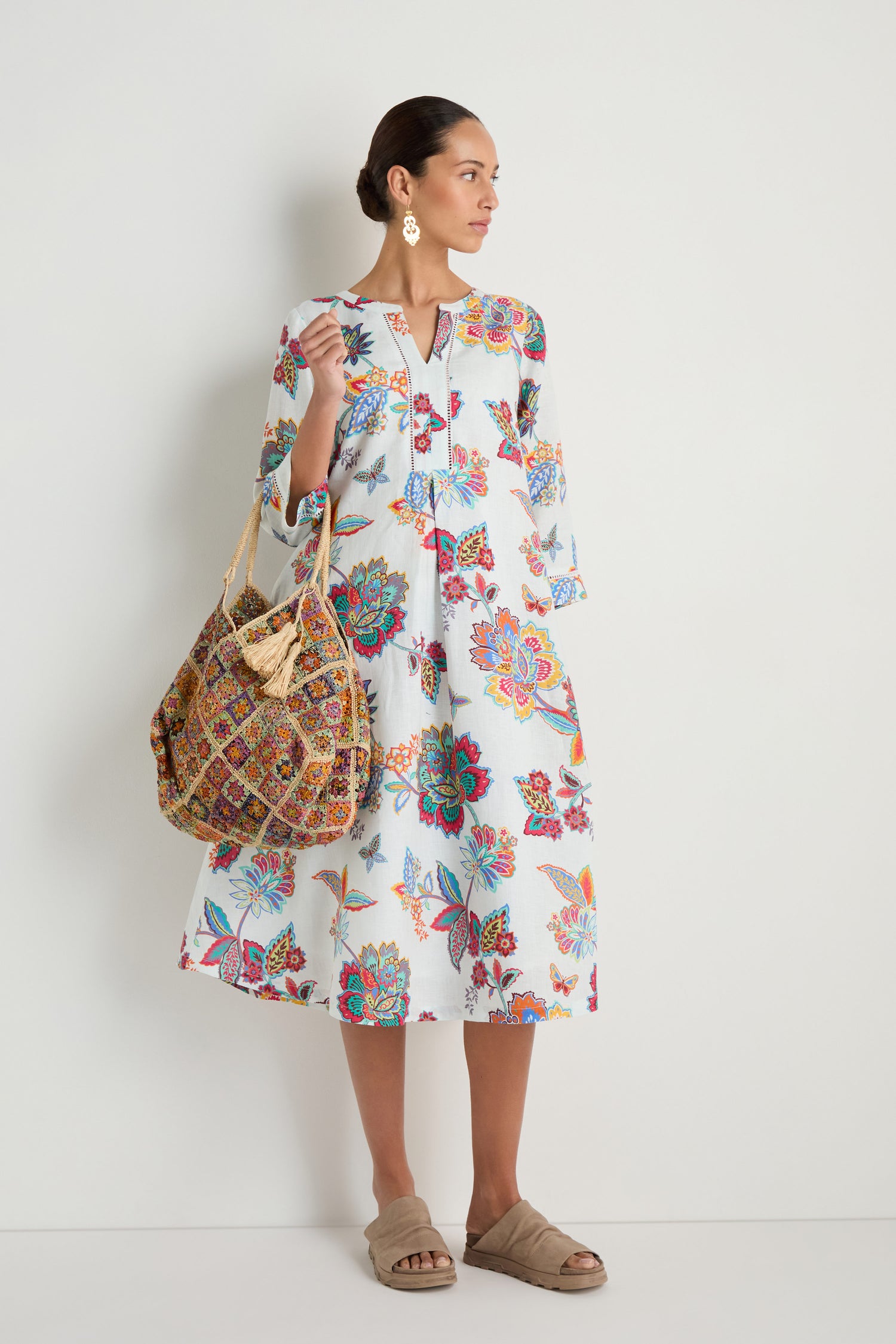 A woman stands against a plain wall in a white floral dress, accessorized with statement earrings, beige slide sandals, and the Handwoven Flower Raffia Bag crafted by Madagascar artisans.