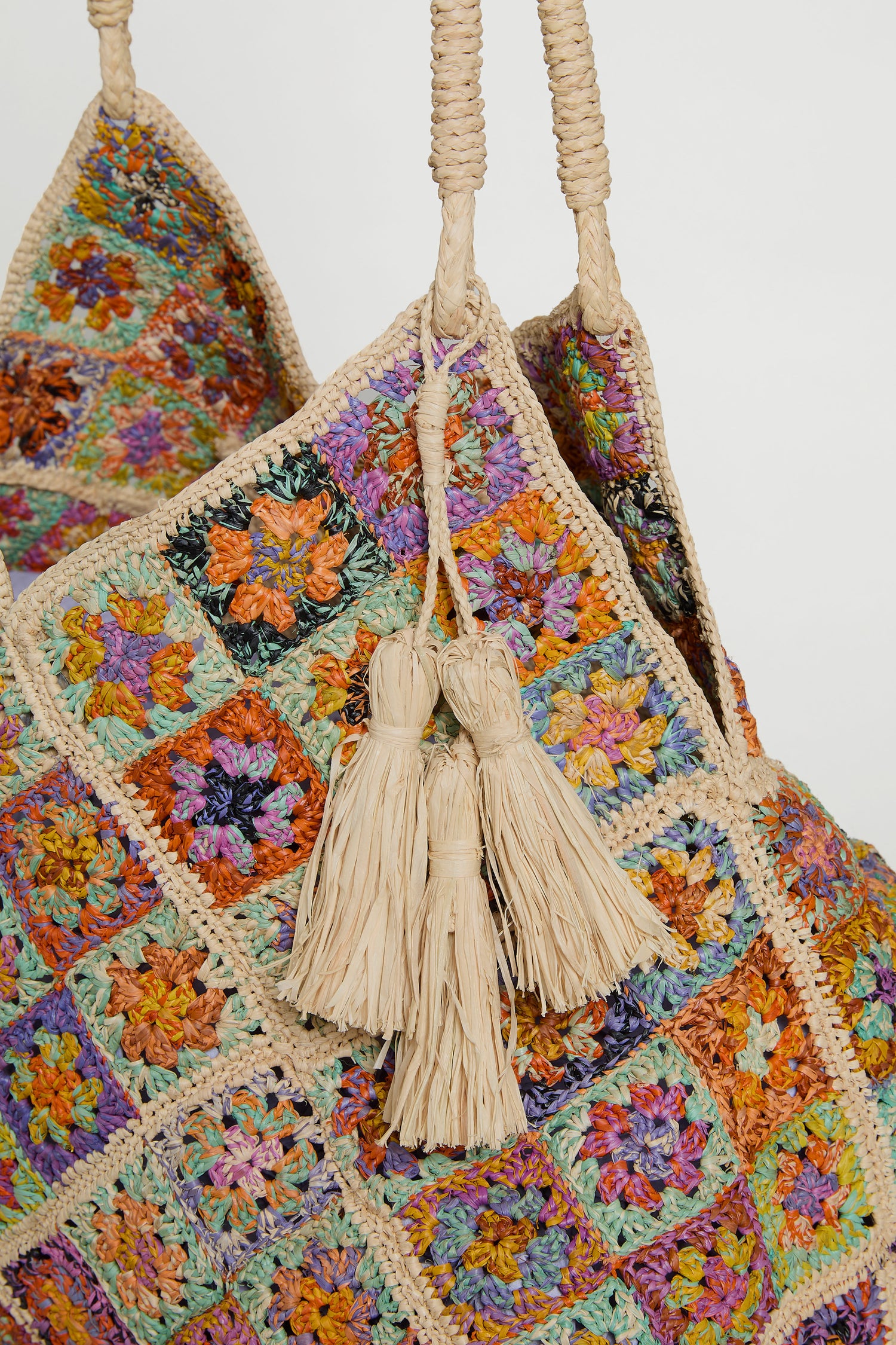 Close-up of the Handwoven Flower Raffia Bag, featuring a colorful patchwork pattern, braided handles with two beige tassels, and floral designs inspired by Madagascar artisans.