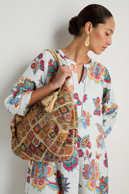 A woman in a floral white dress and gold earrings holds the Handwoven Flower Raffia Bag, a large, multicolored raffia bag featuring vibrant floral patterns crafted by artisans in Madagascar.