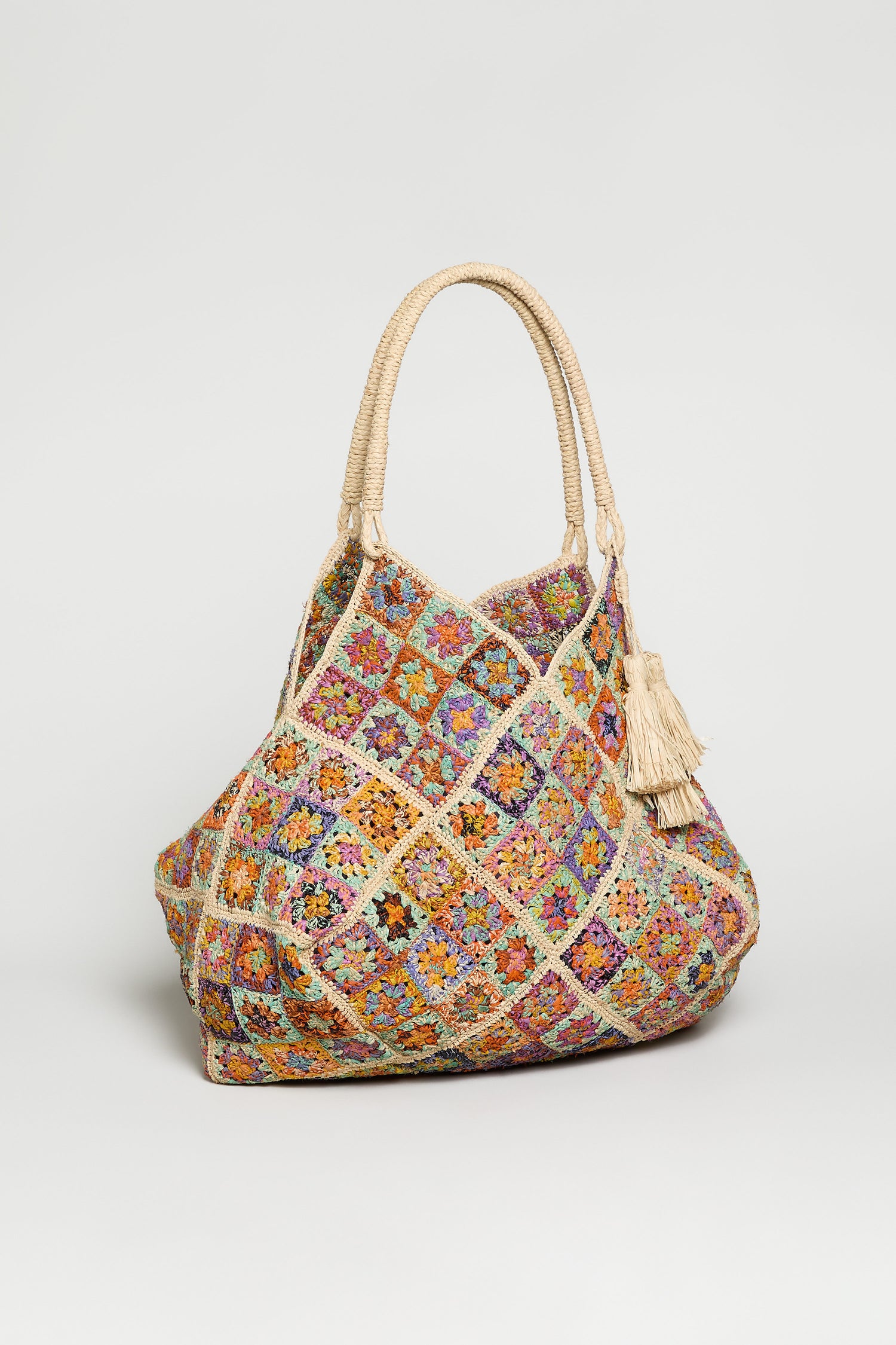 The Handwoven Flower Raffia Bag features a colorful crocheted patchwork, braided rope handles, floral woven designs, and two fringed tassels, all crafted by Madagascar artisans.