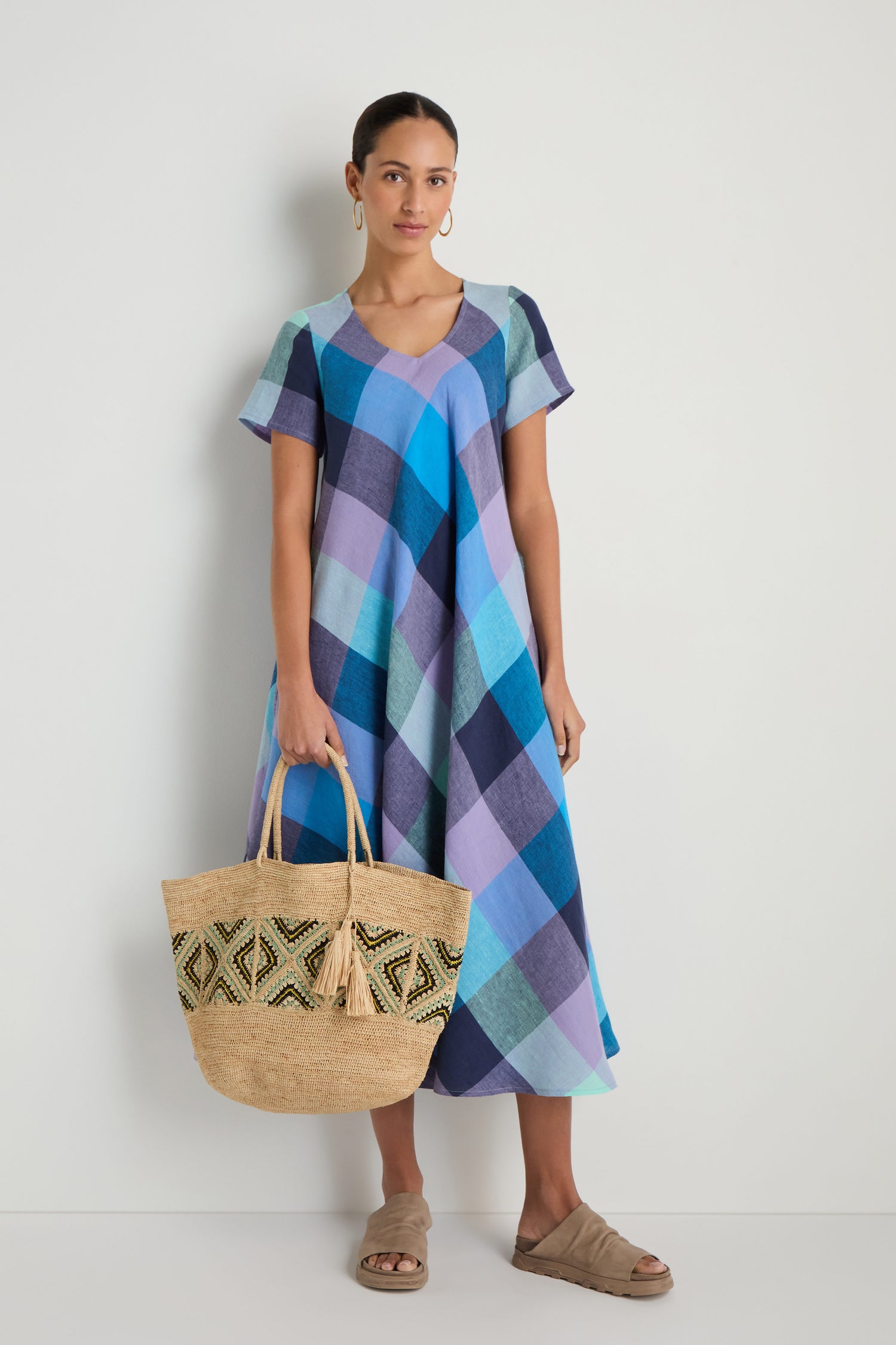 A woman stands against a white wall wearing a short-sleeve blue plaid midi dress, tan sandals, large hoop earrings, and carrying the Handwoven Raffia Tote Bag for a chic and effortless summer look.