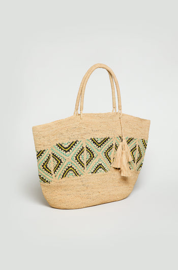 The Handwoven Raffia Tote Bag features dual handles, a green and black geometric pattern, and two decorative tassels on one handle, all set against a plain background.