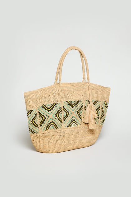 The Handwoven Raffia Tote Bag features dual handles, a green and black geometric pattern, and two decorative tassels on one handle, all set against a plain background.