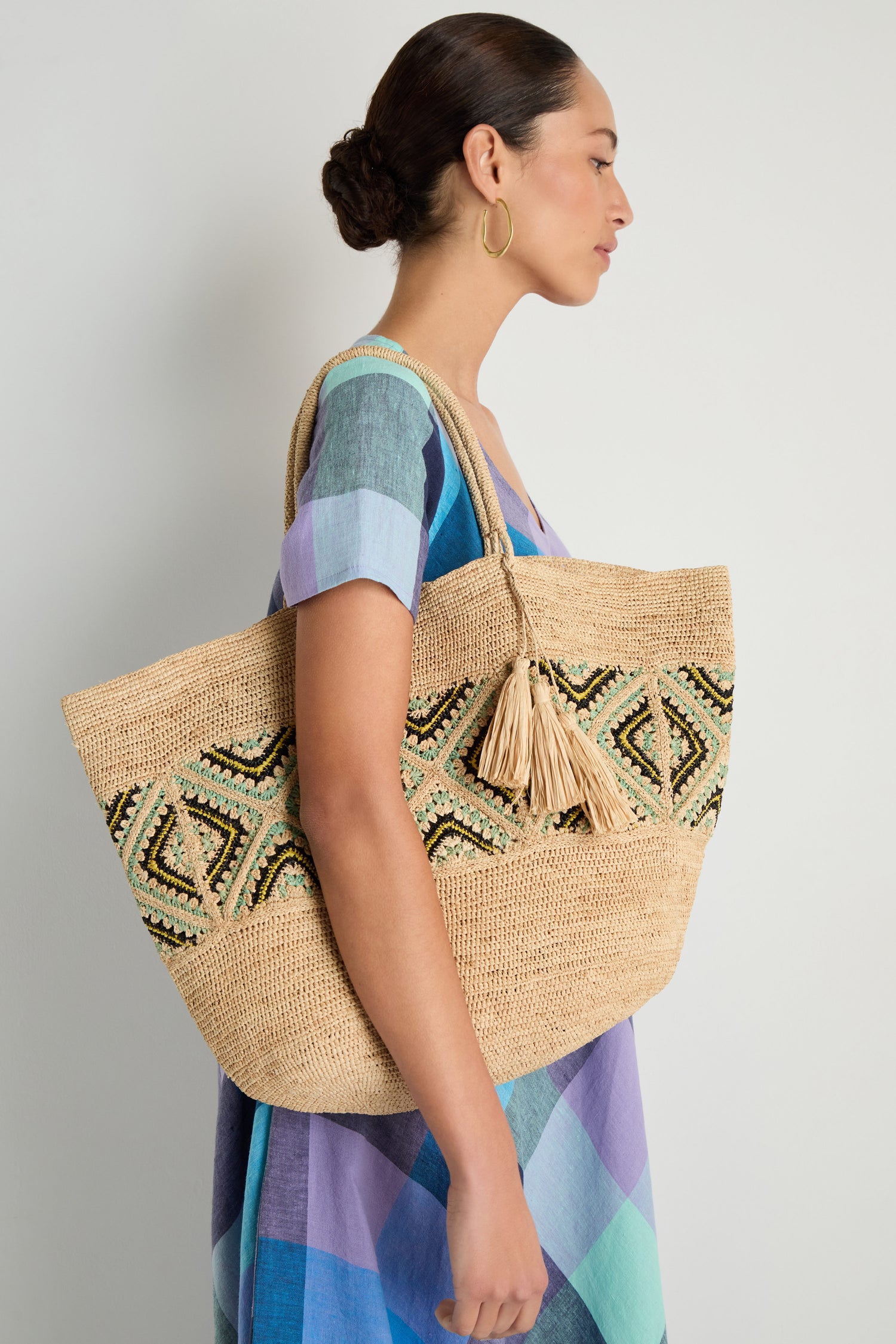 A woman in a colorful checkered dress stands sideways holding the Handwoven Raffia Tote Bag, which features bold geometric patterns and playful tassel details.