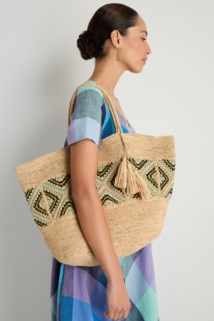 A woman in a colorful checkered dress stands sideways holding the Handwoven Raffia Tote Bag, which features bold geometric patterns and playful tassel details.