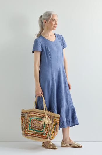 A woman with gray hair in a ponytail wears a knee-length blue dress and gold flats, carrying the Handwoven Crochet Raffia Bag featuring a colorful geometric pattern and tassel.