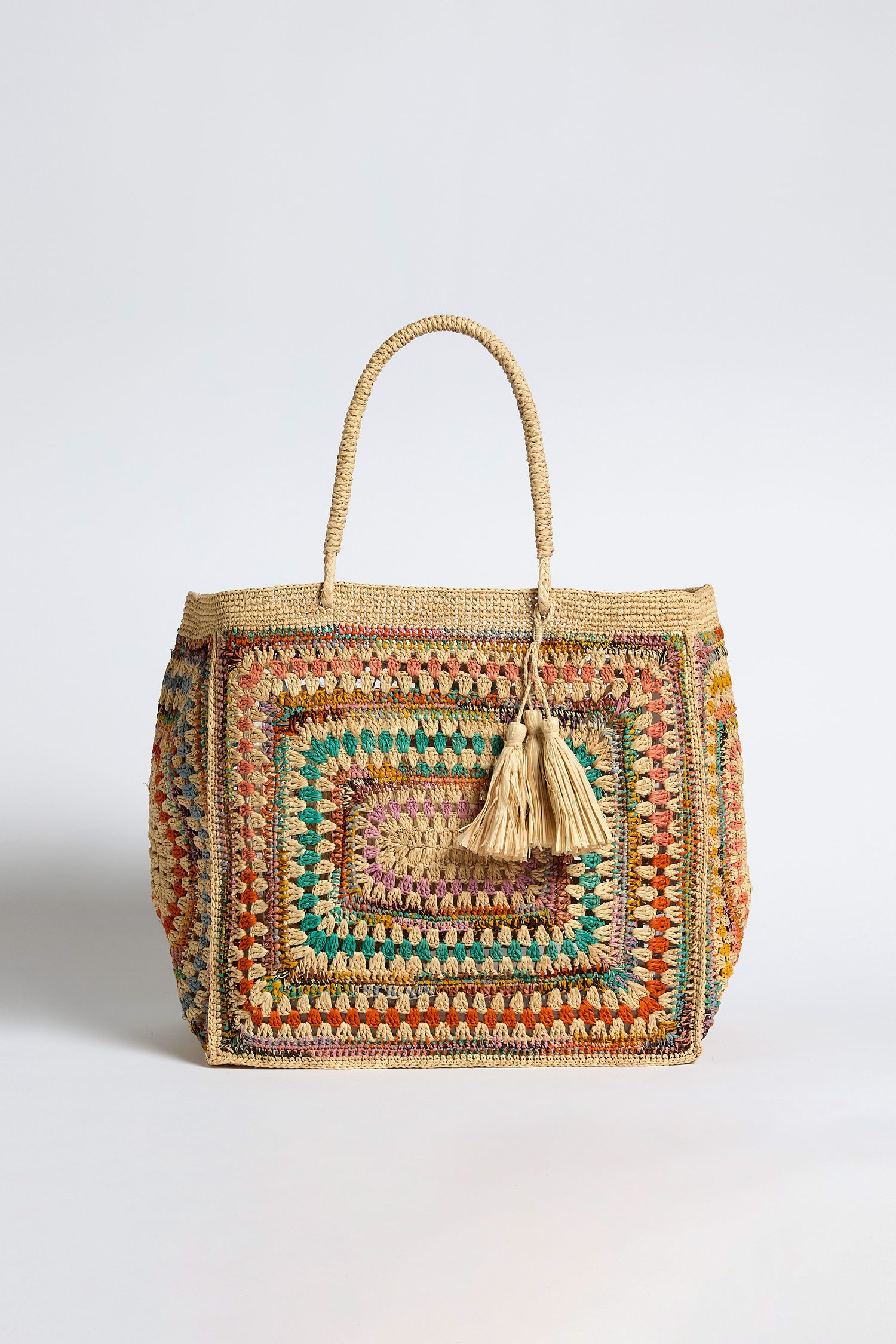 The Handwoven Crochet Raffia Bag is a rectangular artisan handbag with a colorful pattern, two braided handles, and two beige tassels hanging from one handle, displayed against a plain background.