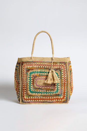 The Handwoven Crochet Raffia Bag is a rectangular artisan handbag with a colorful pattern, two braided handles, and two beige tassels hanging from one handle, displayed against a plain background.