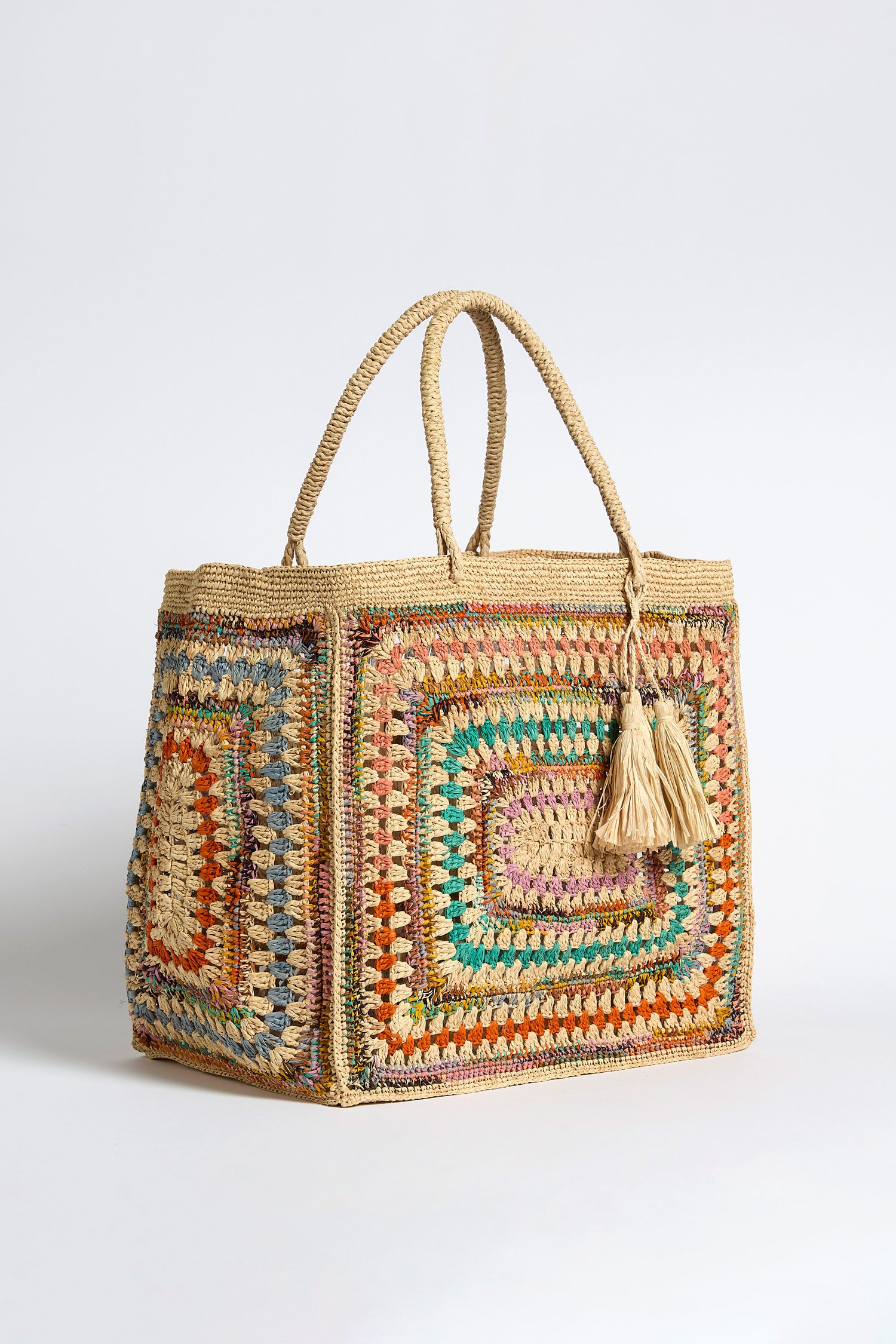 The Handwoven Crochet Raffia Bag features a square shape, colorful crochet pattern, dual rope handles, and two beige tassels, displayed against a plain white background.