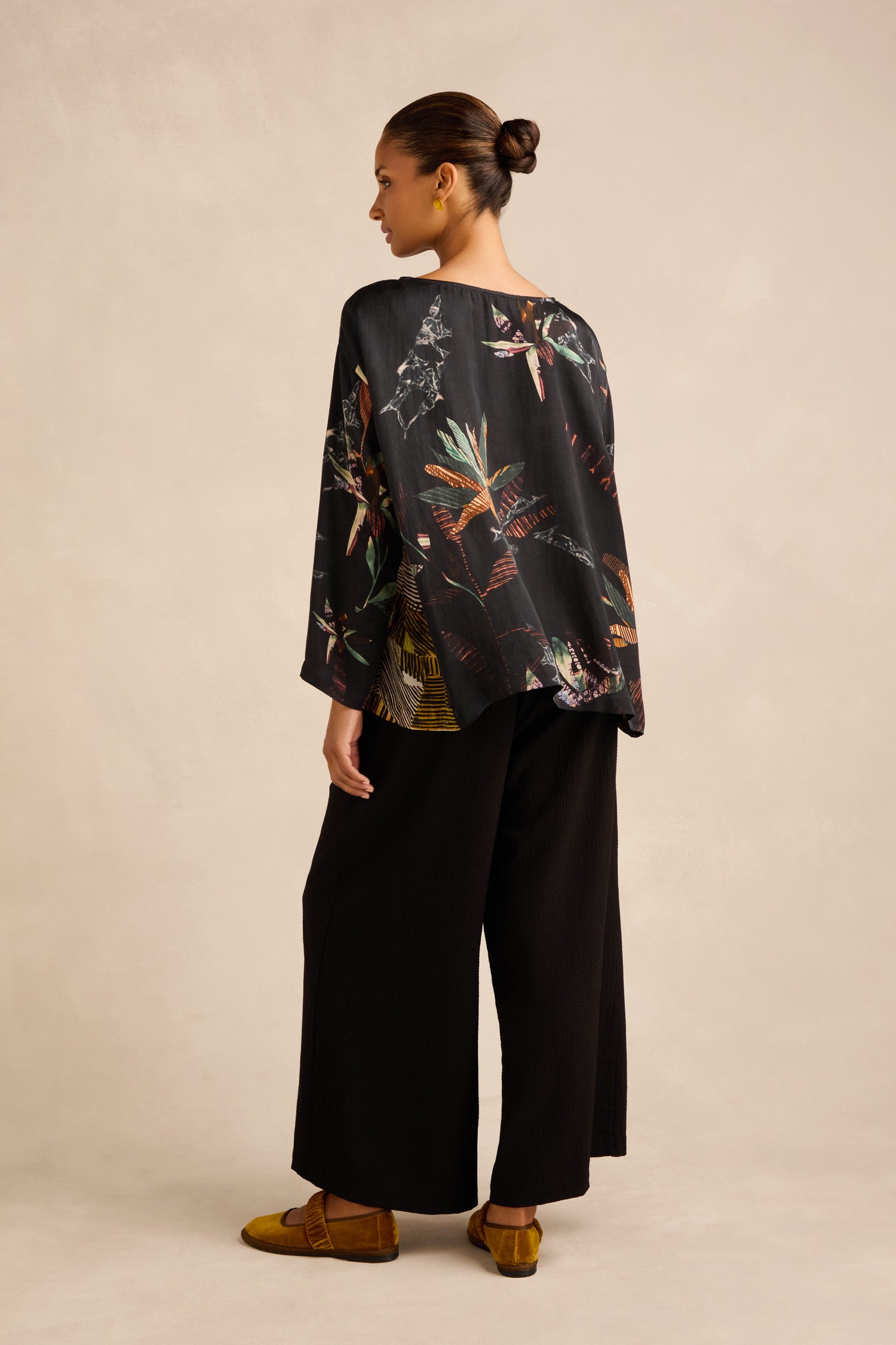 A woman stands facing away, wearing the Abstract Floral Boxy Top in a kimono style, paired with wide black pants and mustard yellow shoes; her hair is in a bun against a neutral background.