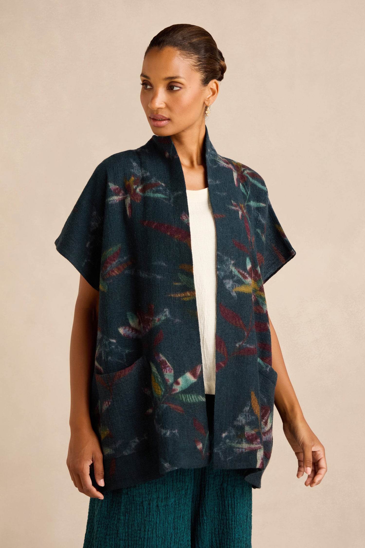 A woman wearing the Abstract Floral Wool Gilet— a dark, open-front cardigan with short sleeves, a shawl collar, and a colorful abstract pattern— layered over a white top and green textured skirt.