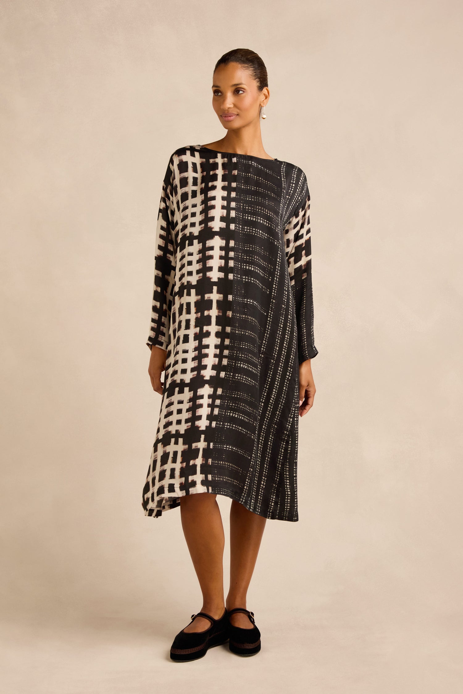 A woman wearing the Check and Dash Easy Dress—a loose-fitting monochrome A-line dress with white grid patterns—paired with black flat shoes and gold hoop earrings, stands against a beige background.