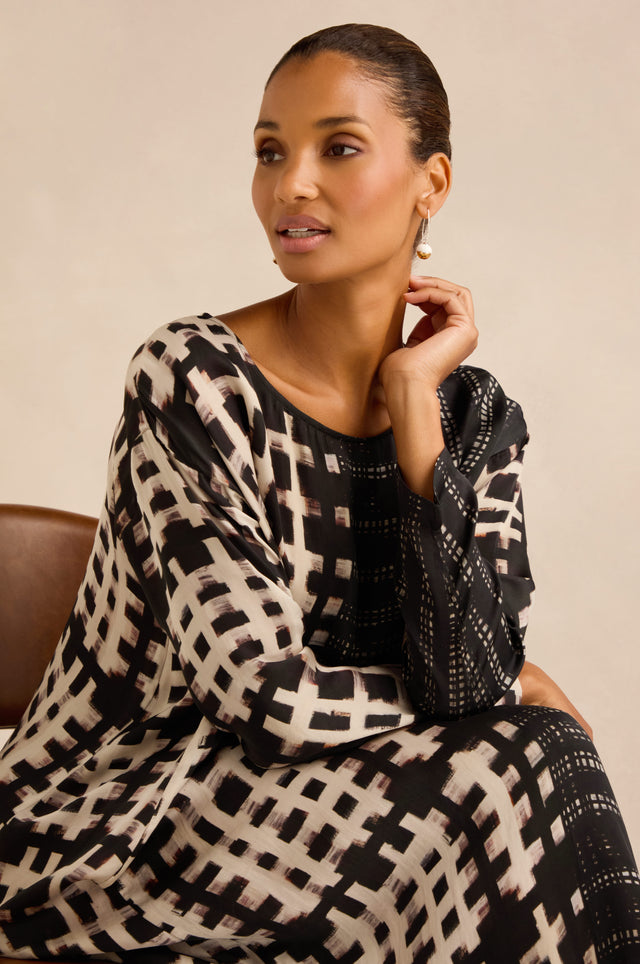 A woman with pulled-back hair sits on a brown chair, resting her hand near her chin and gazing slightly to the side, wearing the Check and Dash Easy Dress—a long-sleeve black and beige patterned cupro dress featuring an elegant A-line silhouette.